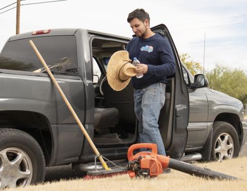 A man in a blue long-sleeved shirt and jeans, holding a shade hat and standing next to a gray truck, with landscaping tools sitting on the ground.