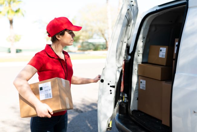 una trabajadora con una camisa roja y un sombrero del mismo color sosteniendo una caja cerrando la puerta trasera de su camión de trabajo 