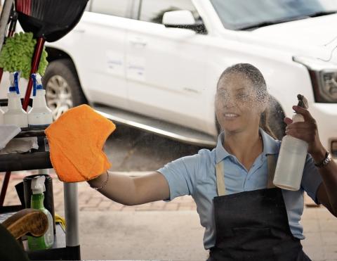 Mujer con un delantal, sosteniendo una botella atomizadora en una mano y limpiando vidrio con la otra mano con un trapo anaranjado.